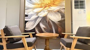 Une pièce avec un grand tableau représentant des motifs floraux, deux chaises en bois, une petite table ronde et un appareil de climatisation.