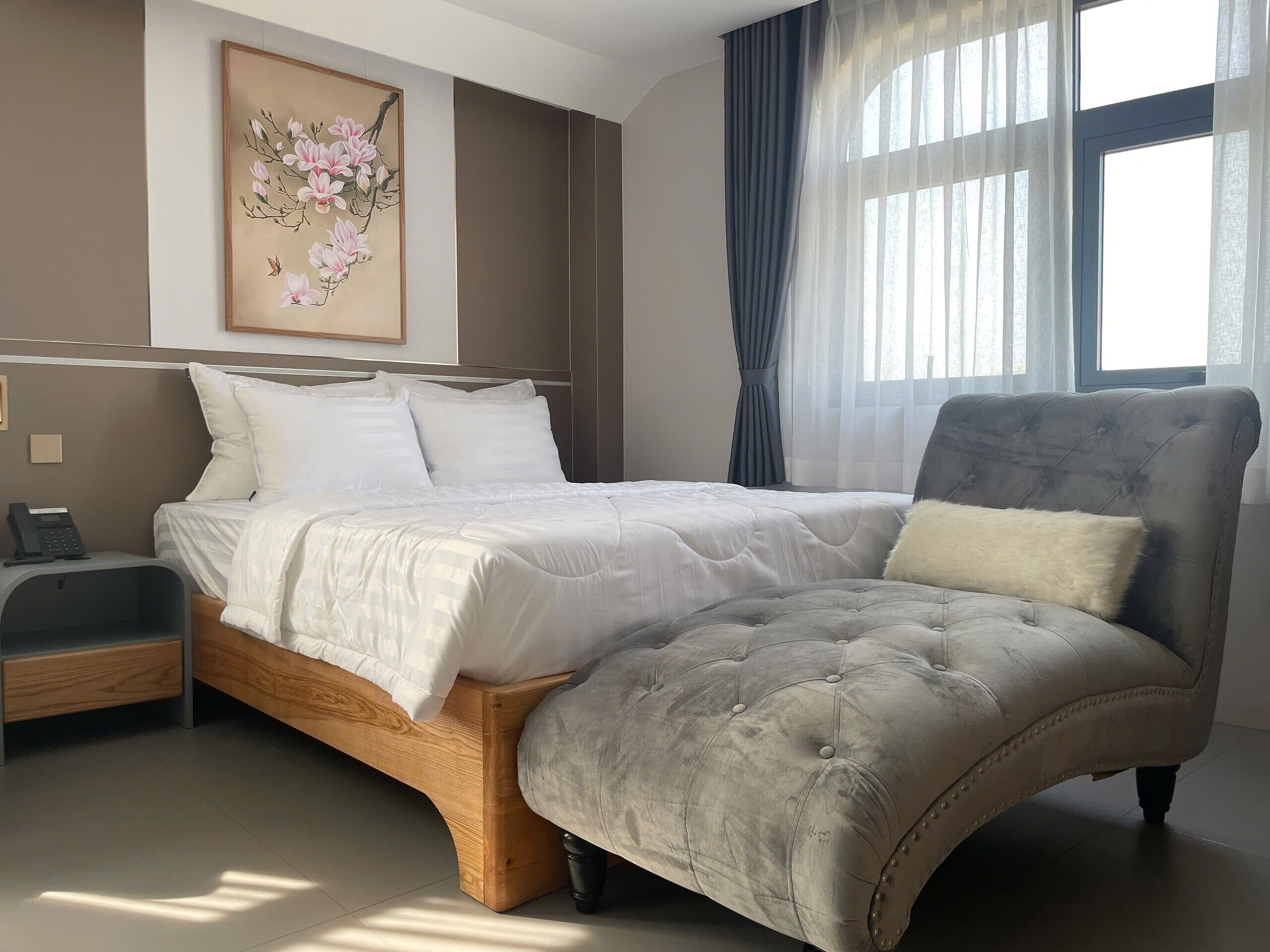 Un dormitorio con cama, mesita de noche, una silla, ventana con cortinas y un cuadro en la pared.