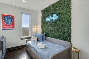 Ein Schlafzimmer mit Bett, Fenster, einer Neonleuchte und einer Wandpflanze.