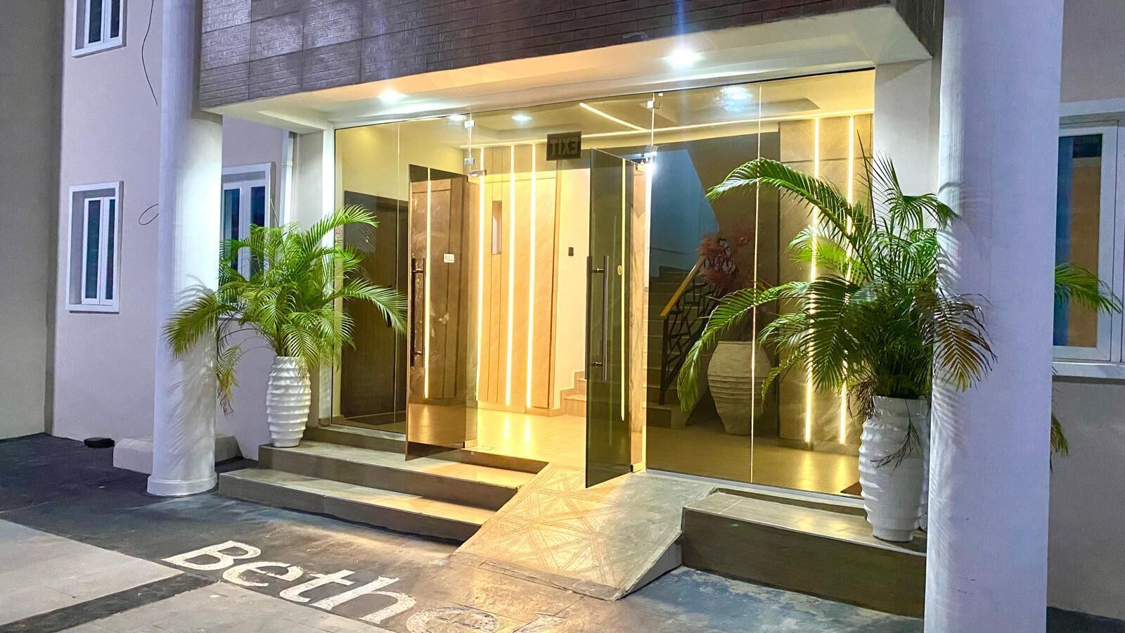 Entrada de un hotel con puerta de vidrio, escalones que conducen a ella y plantas en macetas a ambos lados.