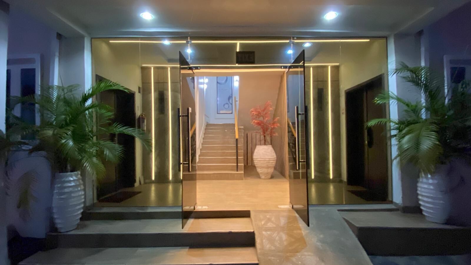 Una entrada de hotel moderna con puerta de cristal, una escalera que sube a los pisos superiores y plantas en macetas a ambos lados.