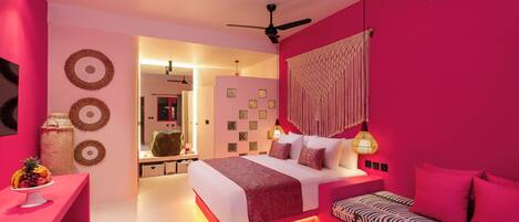 Un dormitorio moderno con una decoración en tonos rosados, una cama con ropa de cama estampada, una mesita de noche con frutero y un ventilador de techo.