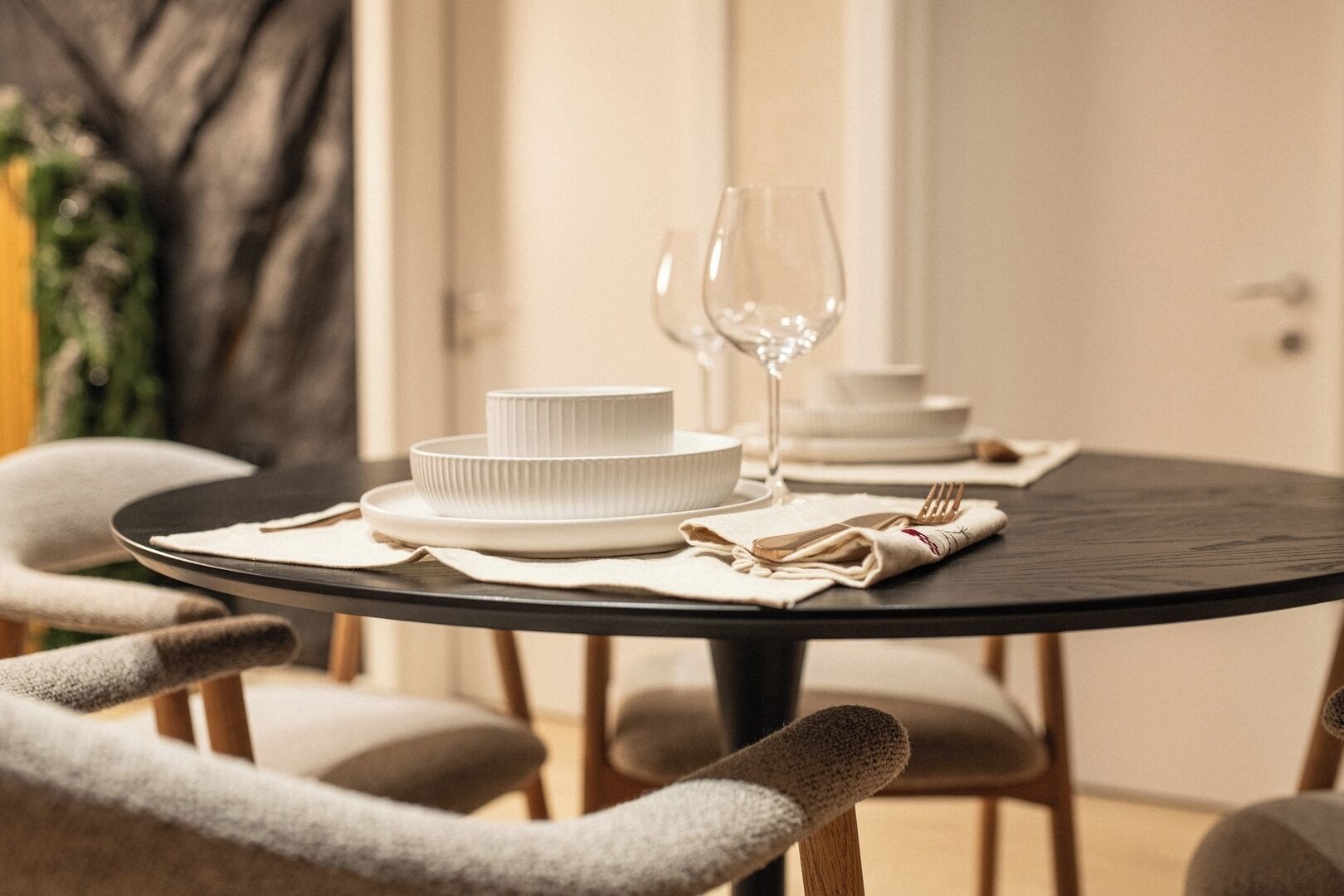 A round dining table with white plates and a wine glass, set with a beige napkin and cutlery.