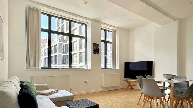 A modern living room with a sofa, a dining table with chairs, a TV, and large windows with a city view. - Shoreditch Apartments by PrimeCityHosts F3 (Greater London)