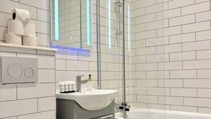 A modern bathroom with white tiled walls, a glass shower enclosure, a white bathtub, and a grey cabinet with a sink. - Shoreditch Apartments by PrimeCityHosts F3 (Greater London)