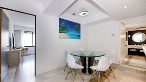 Een moderne eethoek met een glazen tafel en witte stoelen, een schilderij van een strand en een keuken met een ronde spiegel.