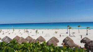 Een strand met rieten parasols, palmbomen en een helderblauwe lucht.