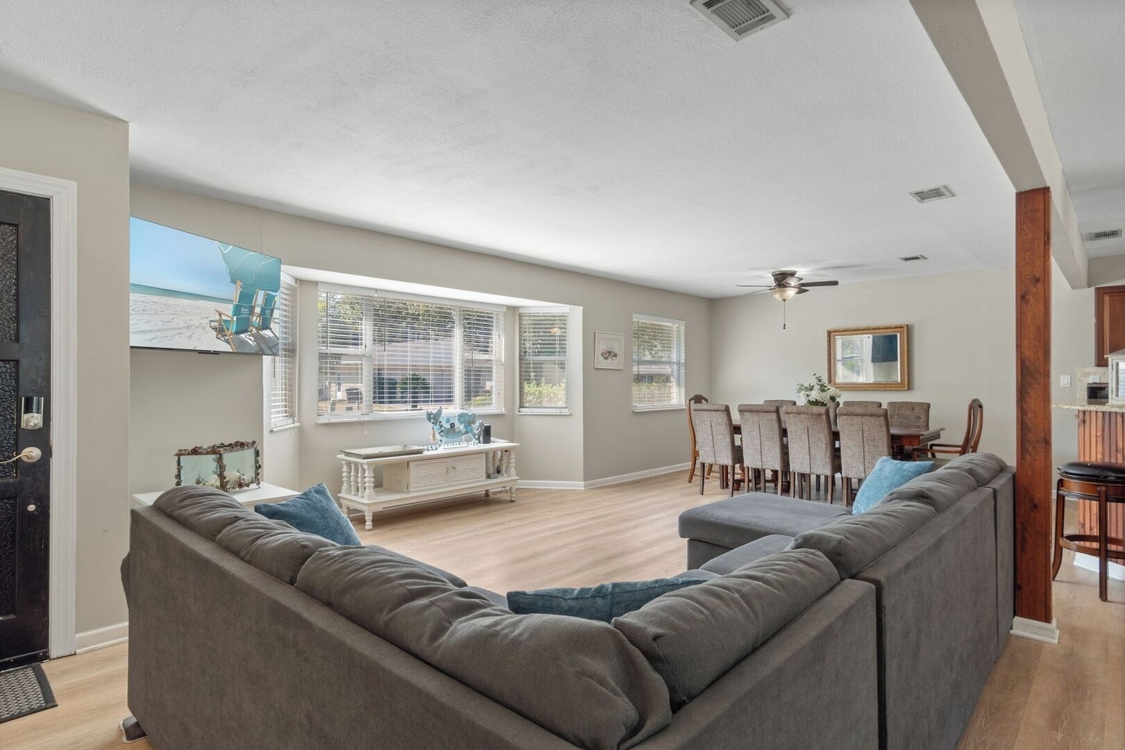 Un salón amplio con un sofá seccional gris, una zona de comedor con mesa y sillas de madera, y ventanas grandes con persianas.