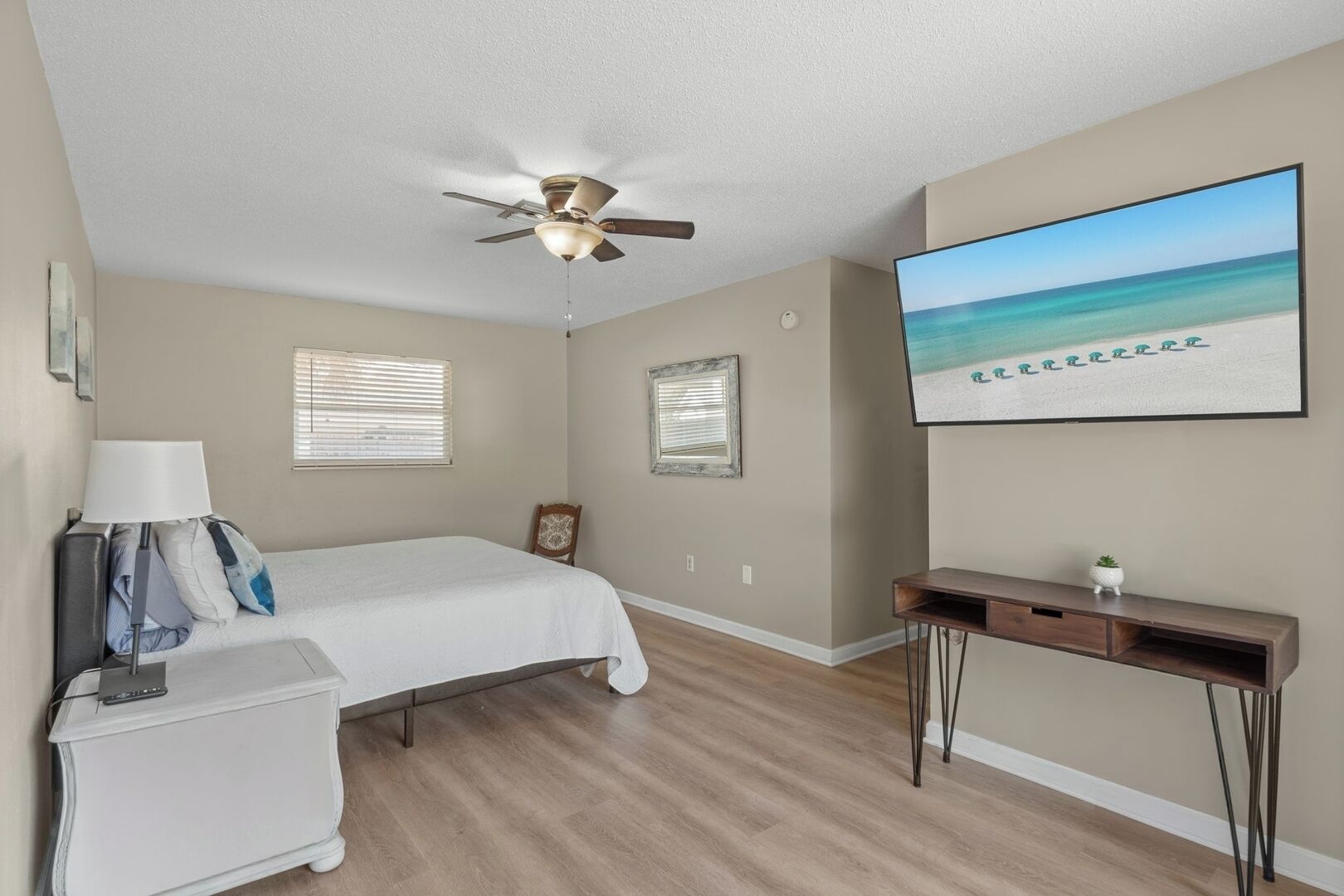 A bedroom with a bed, a desk, a ceiling fan, and a beach-themed painting on the wall.