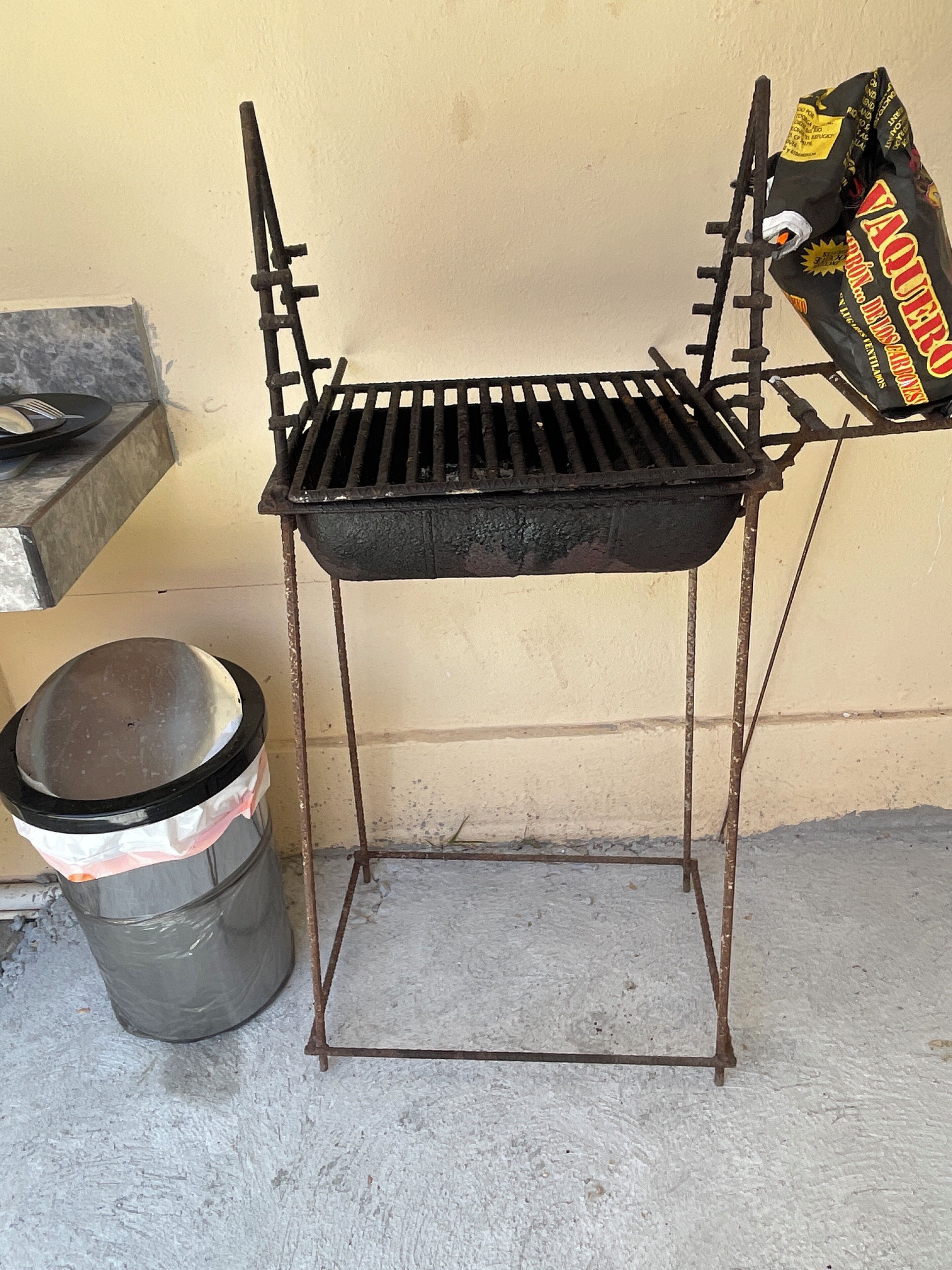Een draagbare grill met een metalen frame en rooster, een prullenbak met deksel, en een zak houtskool in de buurt.
