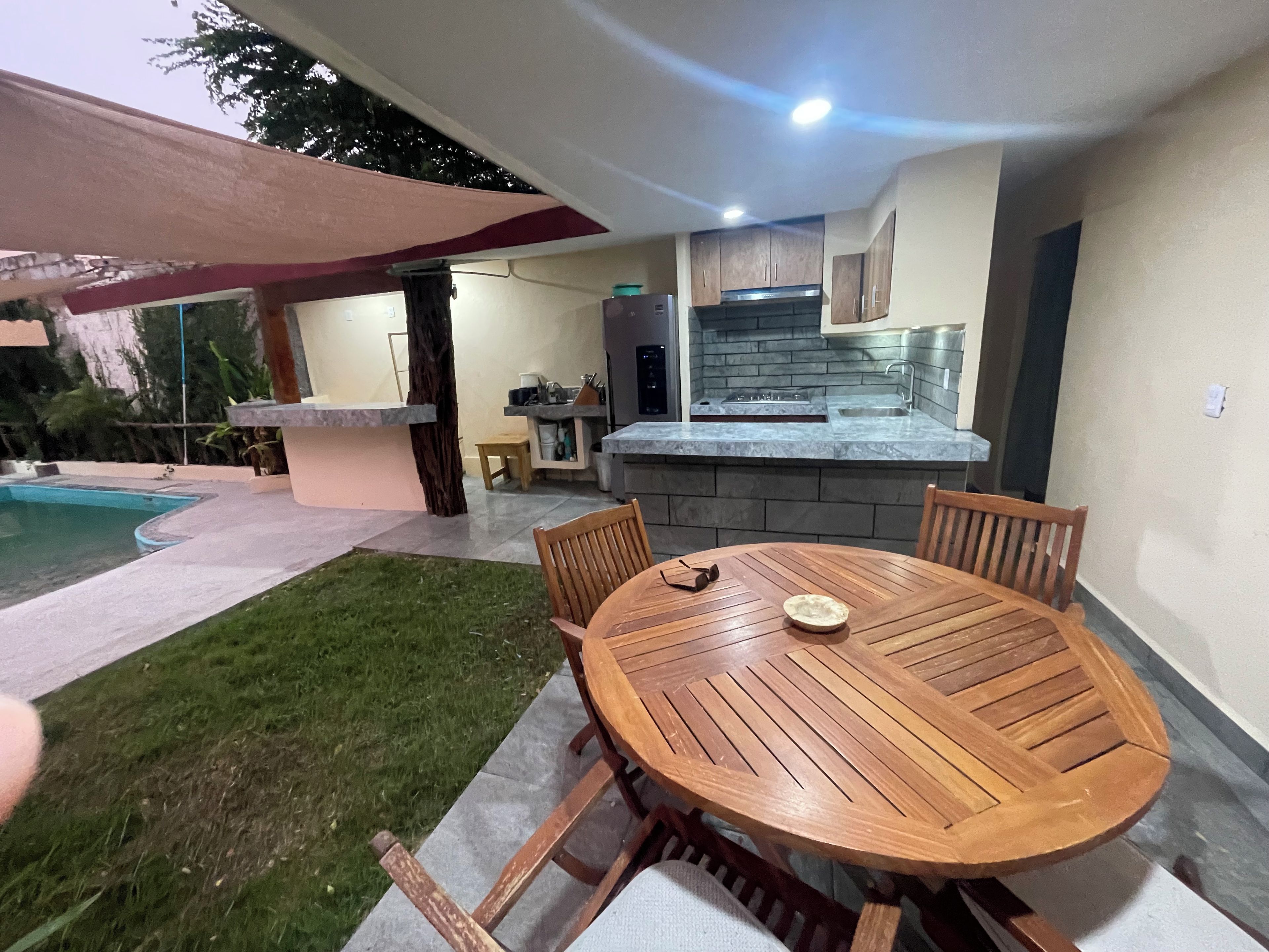 Un espace extérieur aménagé pour les repas, avec une table et des chaises en bois rondes, une piscine et une cuisine dotée de plans de travail en pierre.