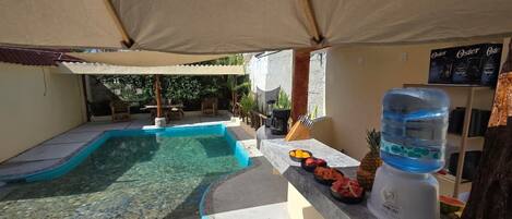 A pool area with a water dispenser, fruits, and a covered seating area.
