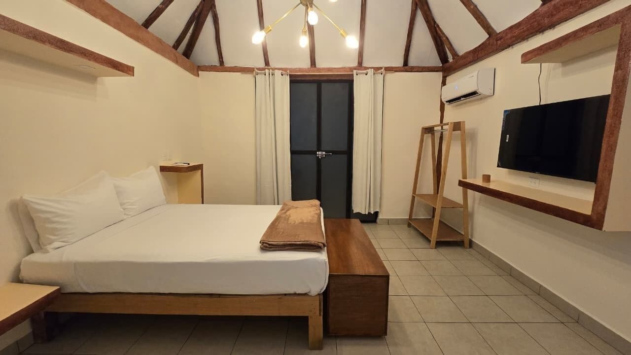 A bedroom with a wooden bed, a bedside table, a bench, a TV mounted on the wall, and a door with curtains.