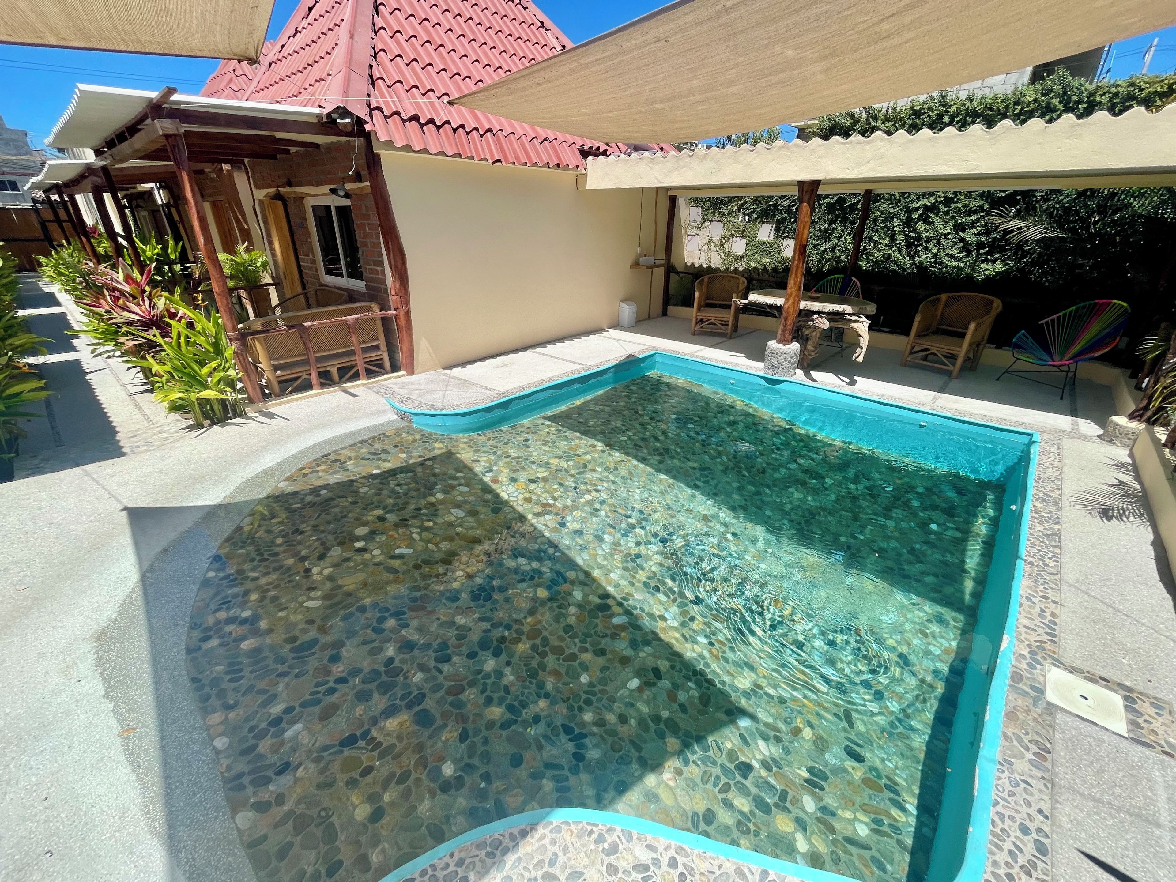 Una piscina cristalina con fondo de piedras, rodeada por un patio con mobiliario exterior y plantas.