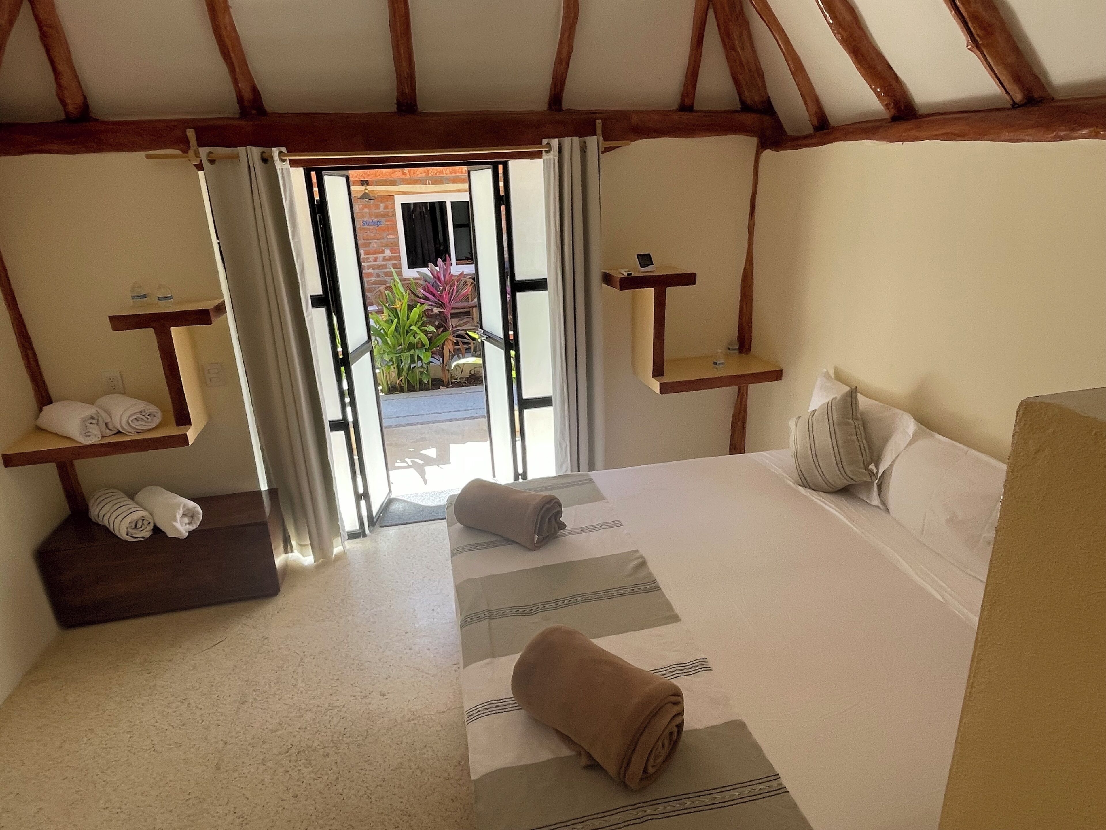 Un dormitorio con una cama, una estantería de madera con toallas, una mesita con un teléfono y vistas al exterior.