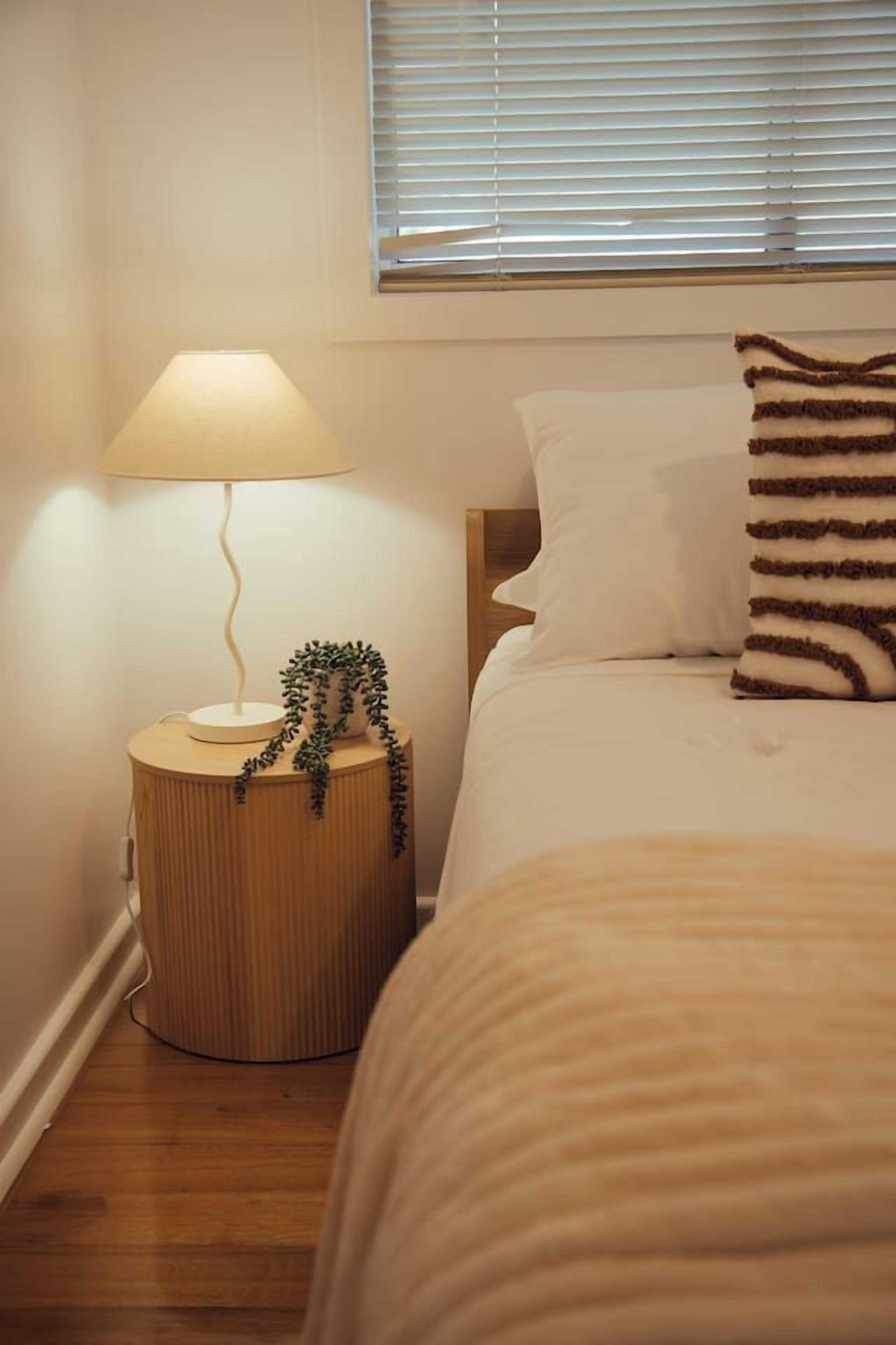 Ein Schlafzimmer mit einem Bett, einer Nachttischlampe, einem kleinen Holfnachtkästchen und einem Fenster mit Jalousien.