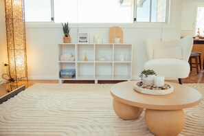 A modern living room with a white sofa, a round wooden coffee table, a white rug, and a white shelving unit with various decorative items. - Modern 3BR Claremont Home | Parking & WiFi (Claremont)