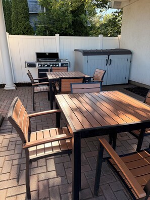 En udendørs terrasse med grill, træbord og stole.