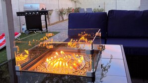 Een terras buiten met een tafel met glazen blad, een barbecue en een blauwe bank.