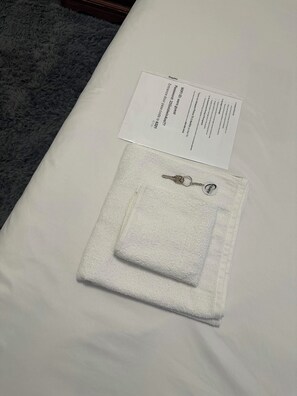 Un lit bien fait, avec une serviette blanche pliée et un porte-clés avec une clé. On peut également voir un papier sur lequel est écrit un texte et un numéro.