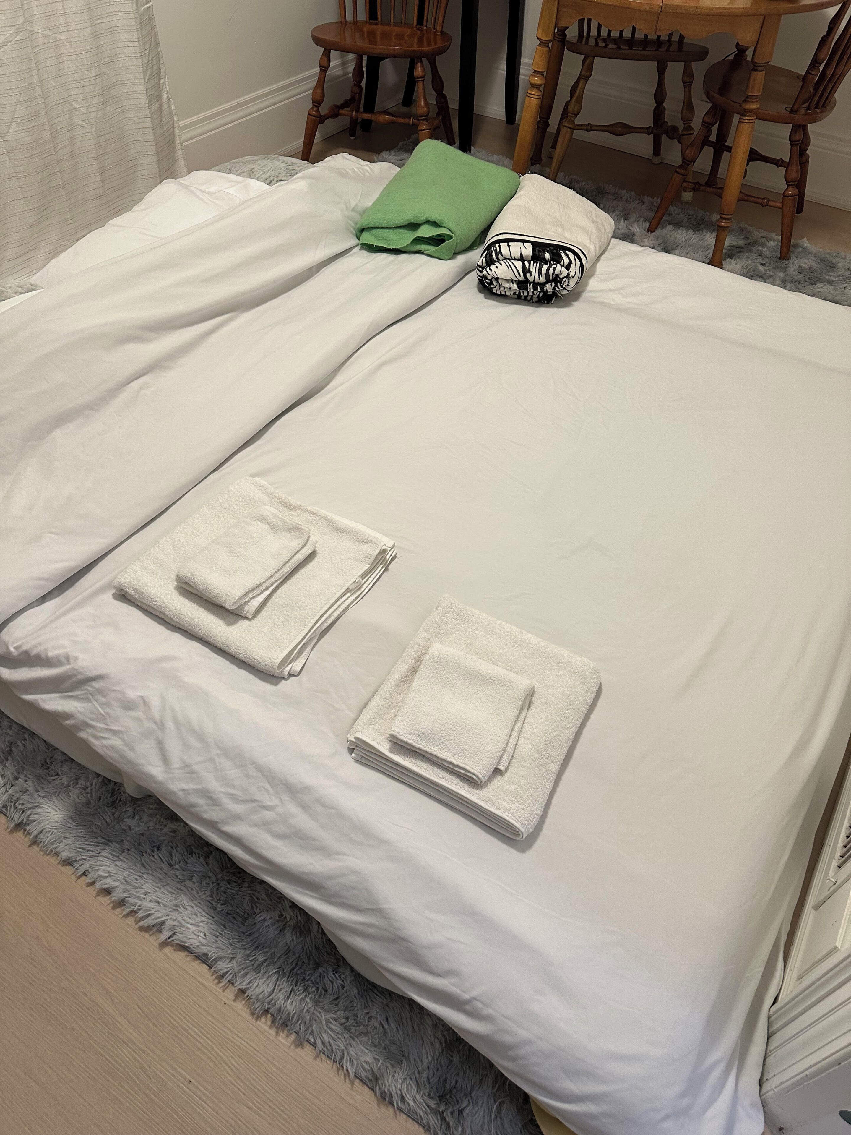 A neatly made bed with white linens, two folded towels, a green towel, and a black and white patterned pillow.