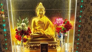 A statue of a seated figure in a golden robe, surrounded by flowers and candles, with a patterned backdrop and decorative lights.
