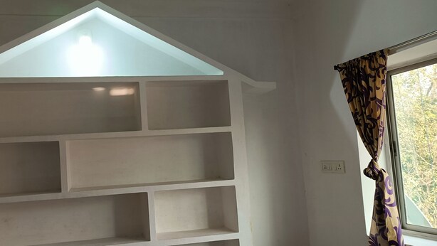 A room with a sloped ceiling, a built-in white shelving unit, a window with patterned curtains, and a visible electrical outlet.