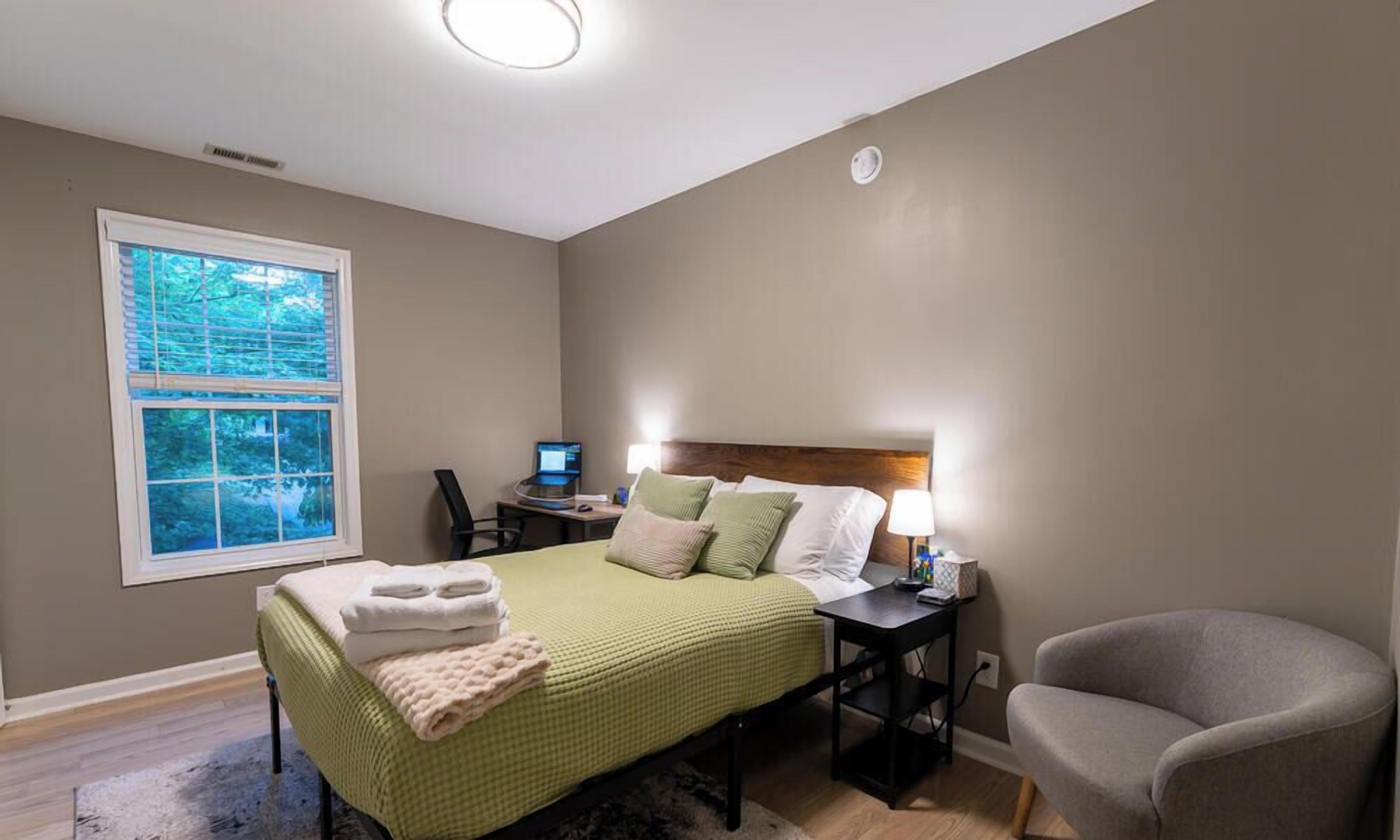 Een slaapkamer met een bed, een bureau, een stoel, een raam en een lamp.