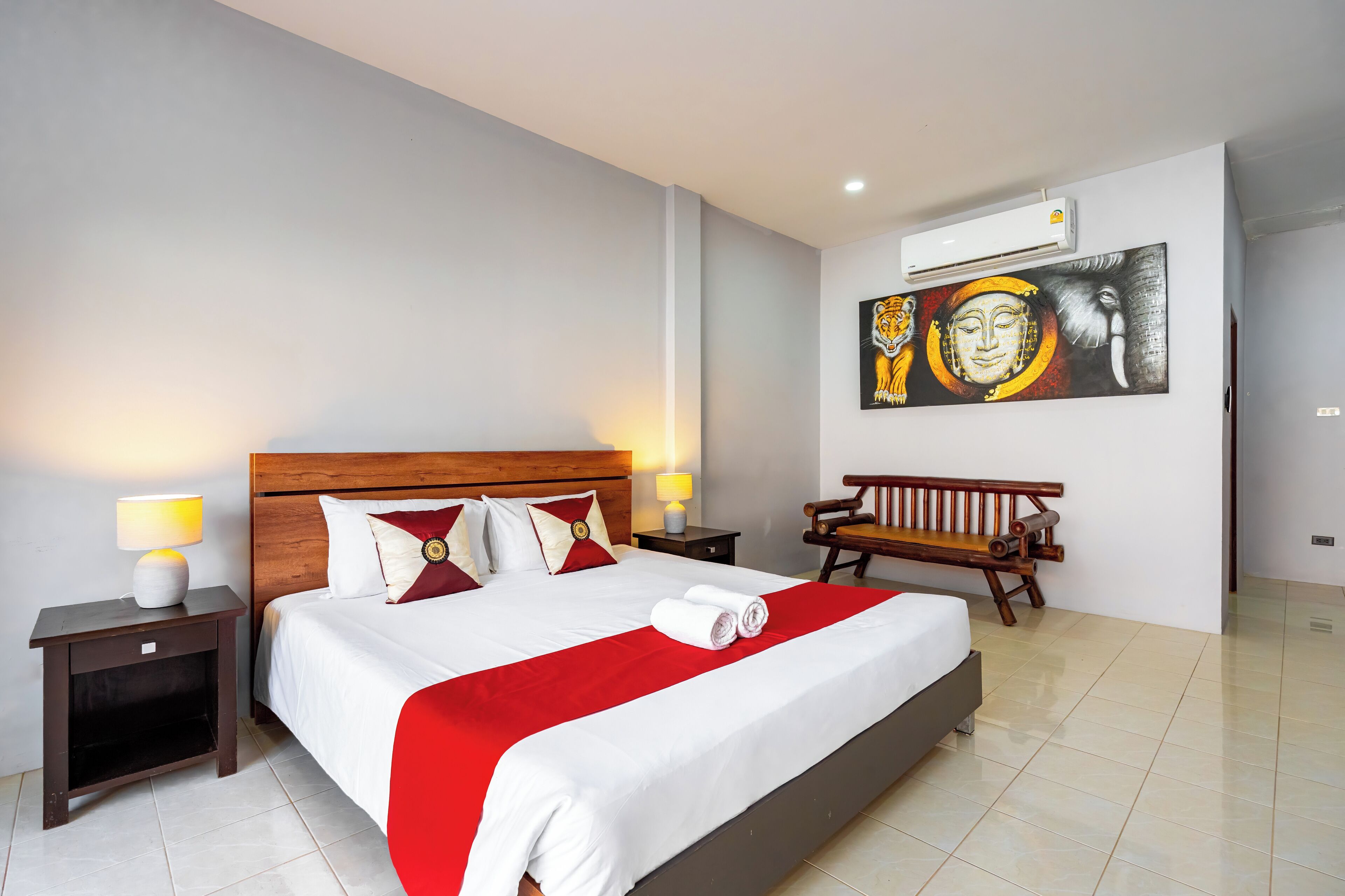 A hotel room with a large bed, a wooden bench, a painting on the wall, and an air conditioning unit.