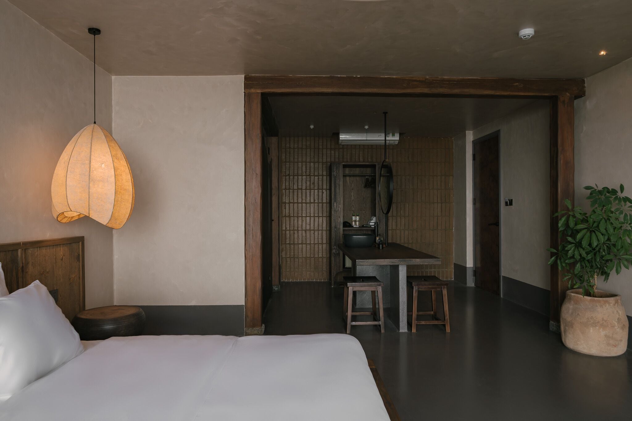 Habitación de hotel con cama, lámpara colgante, un taburete pequeño, planta en maceta y vistas al área de la cocina contigua.