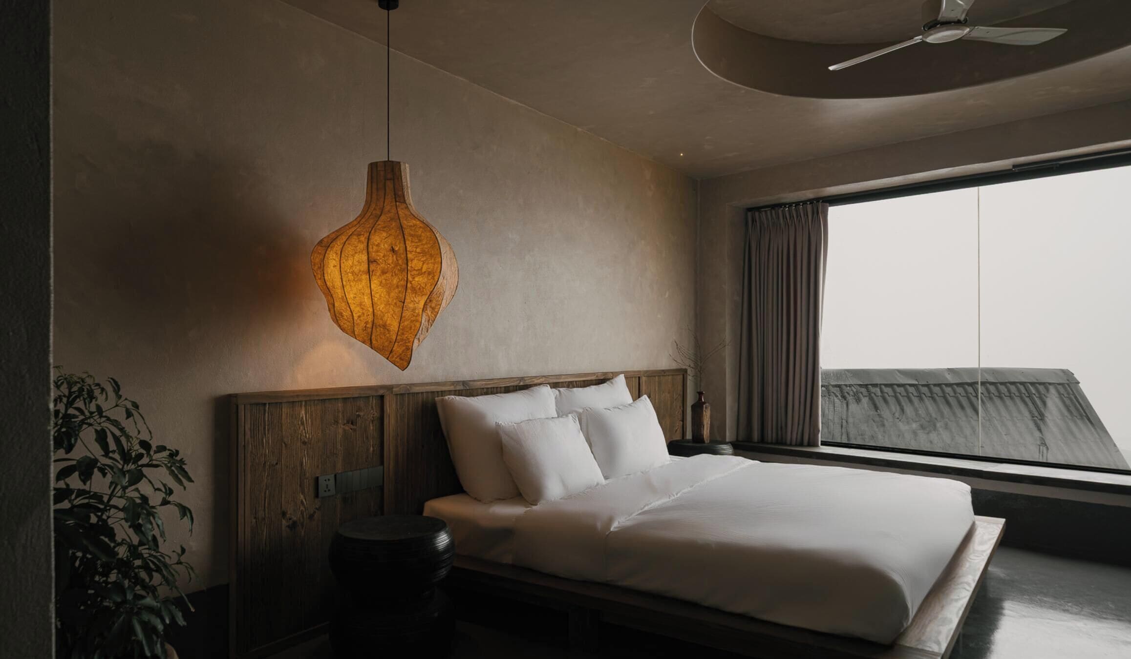 A modern hotel room with a large bed, a hanging light fixture, and a view of the city.