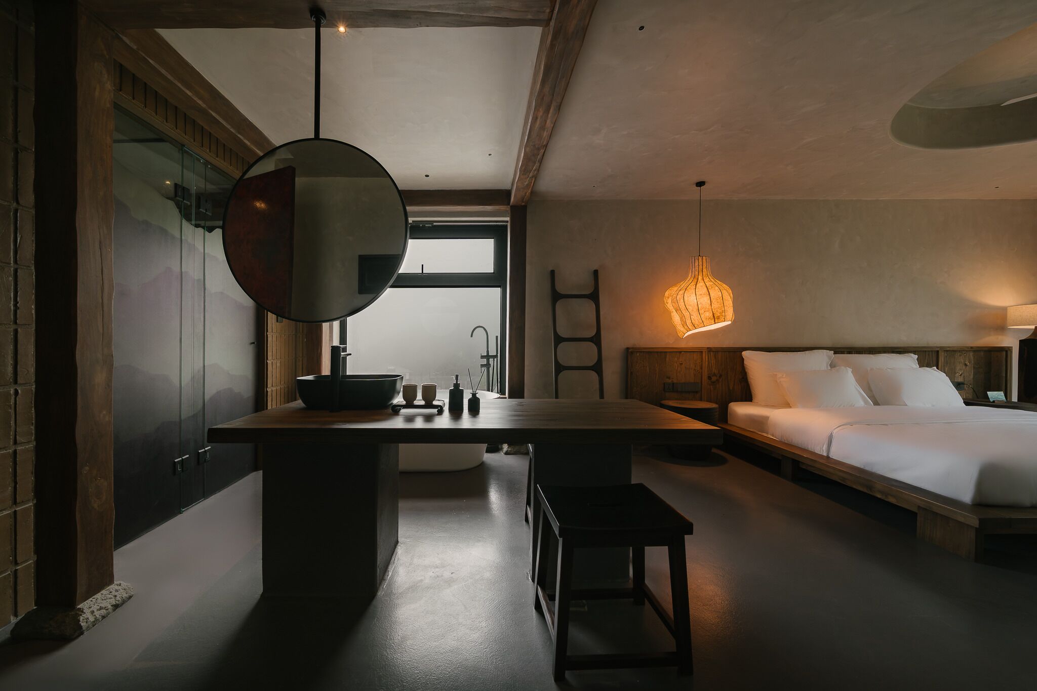 A modern bedroom with a large bed, a wooden nightstand, a round mirror, and a hanging light fixture.