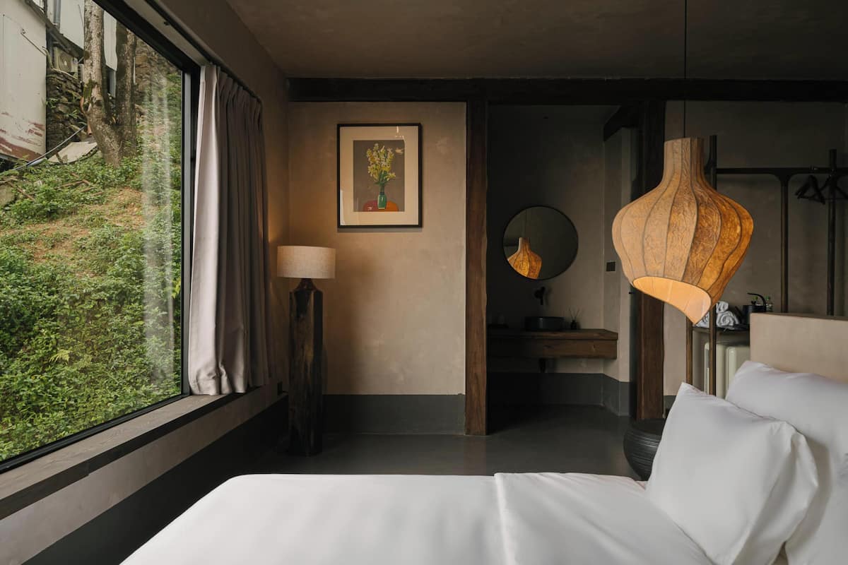 Habitación de hotel con cama, lámpara de noche, un cuadro enmarco, espejo y vista a zonas verdes.