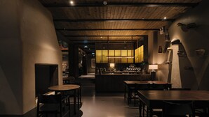 Un interior tenuemente iluminado con vigas de madera en el techo, una zona de bar central y mesas y sillas dispuestas para comer.