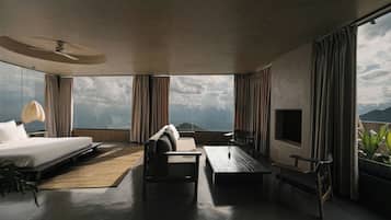 A modern hotel room with a large bed, a sofa, a coffee table, and a TV. The room has large windows offering a view of the ocean and mountains.