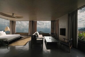 Une chambre d’hôtel moderne dotée d’un grand lit, d’un canapé, d’une table basse et d’un téléviseur. La chambre dispose de grandes fenêtres offrant une vue sur l’océan et les montagnes.