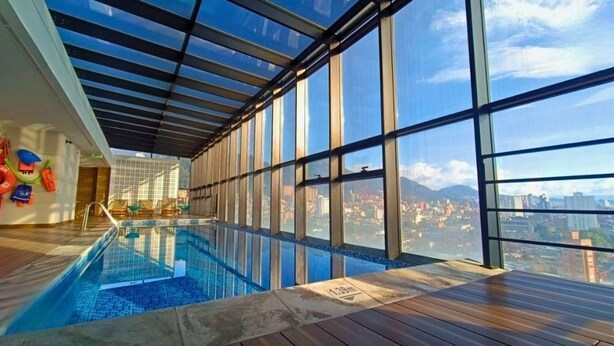 Una piscina interior con vistas a la ciudad, amplias ventanas y una terraza de madera.