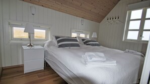 Et soveværelse med skrå væg, en seng med hvide sengetæpper og stribede puder, et natbord med en lampe og et vindue med udsigt.