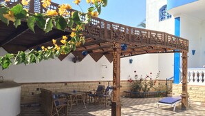 Ein überdachter Patio-Bereich mit Gartenmöbeln, einem Pool und einer Pergola mit gelben Blumen.