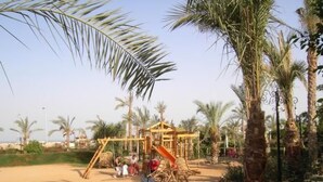 A sandy playground area with a wooden swing set and a slide, surrounded by palm trees and a clear sky. - Fewo Riviera Sharm by Interhome (Sharm El Sheikh)