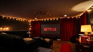 Una camera accogliente con un grande schermo che mostra 'NETFLIX', un divano comodo, un tavolino con una lampada e un tappeto rosso.