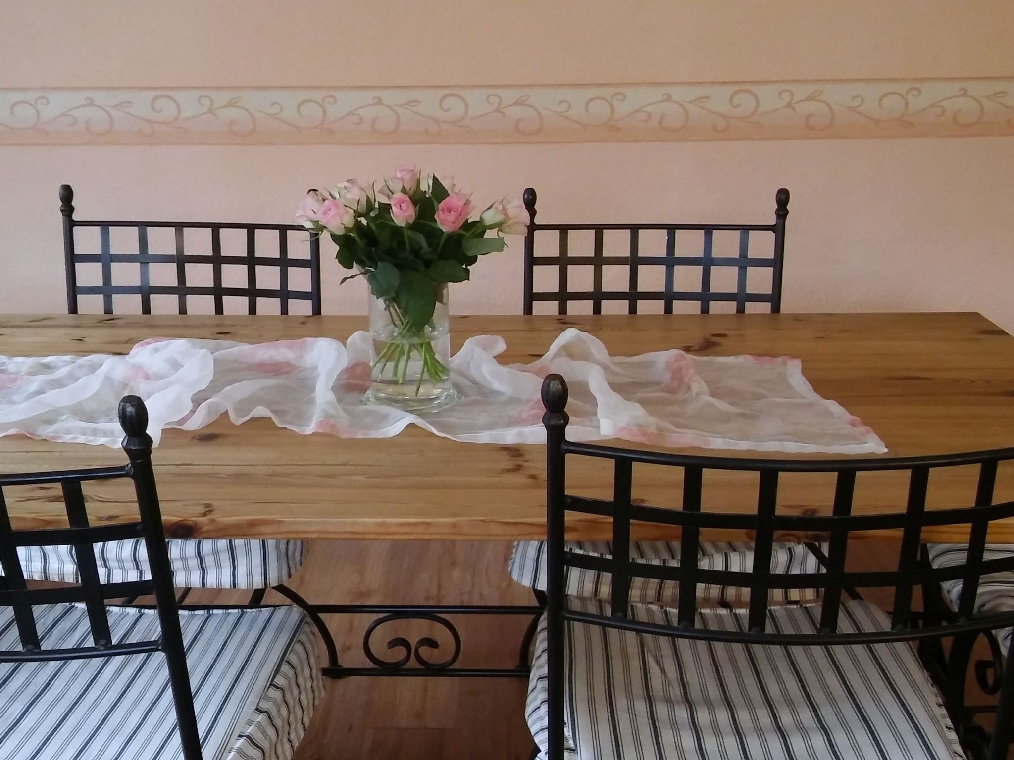 Una mesa de madera para comer con un centro de flores, rodeada por cuatro sillas con estructura de metal negro y cojines a rayas.
