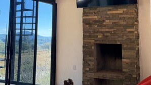 Un salón con chimenea de piedra, un televisor de pantalla plana grande montado encima y un ventanal con vistas al exterior.