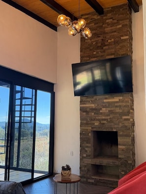 A living room with a stone fireplace, a large flat-screen TV mounted above it, and a large window offering a view of the outdoors.