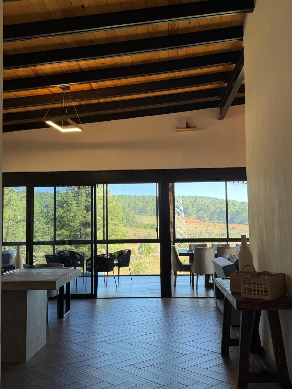 A spacious room with a large window, wooden ceiling beams, and a view of a forested landscape.