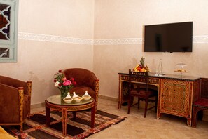 Un salon comprenant un canapé, une table basse ornée d’un vase de fleurs, un téléviseur fixé au mur et une table décorative sur laquelle sont disposés des fruits et un verre.