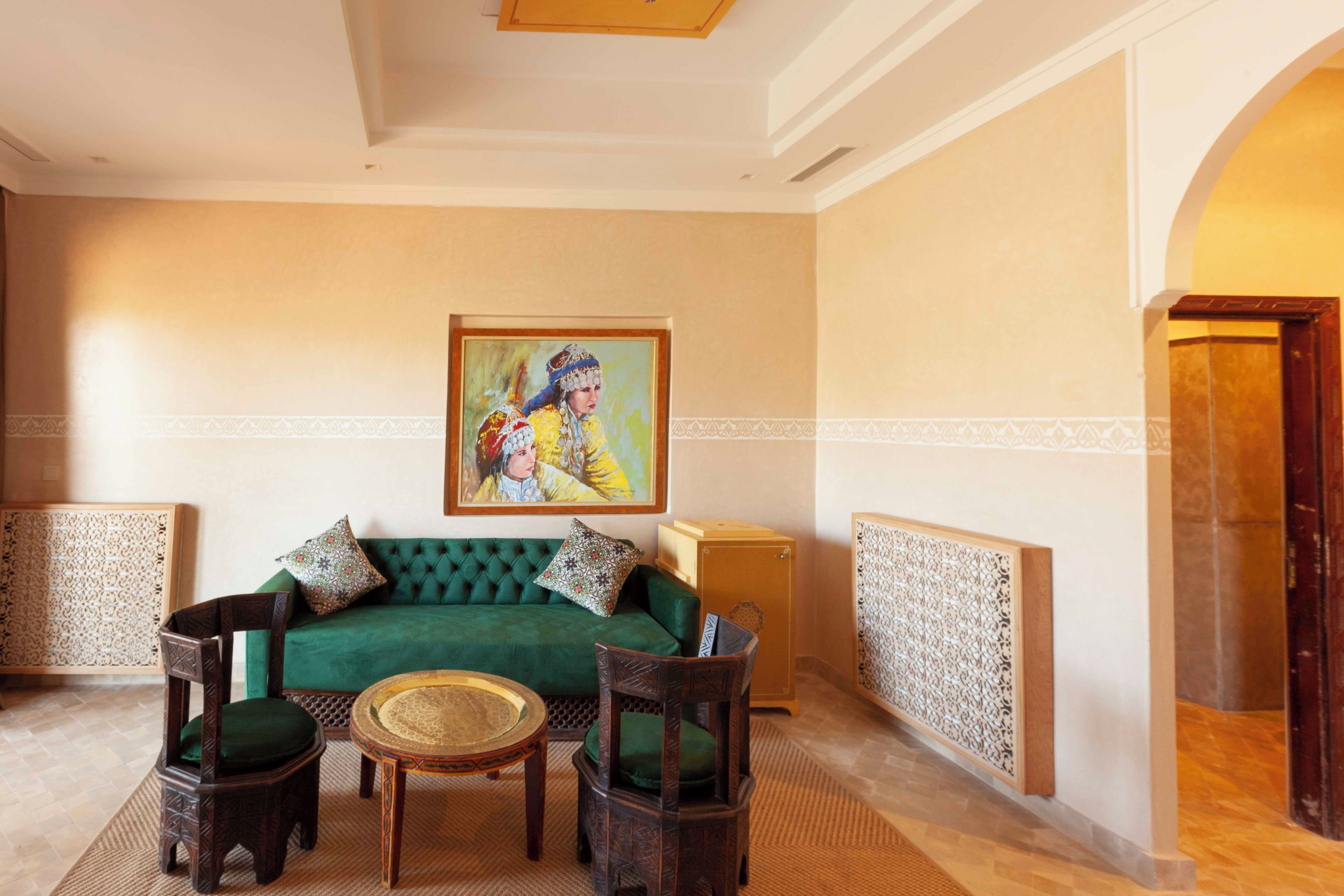 A room with a green sofa, two wooden chairs, a small round table, and a painting on the wall.