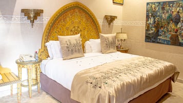 A large bed with ornate headboard, multiple pillows, a bedside table with a lamp, and a painting on the wall.