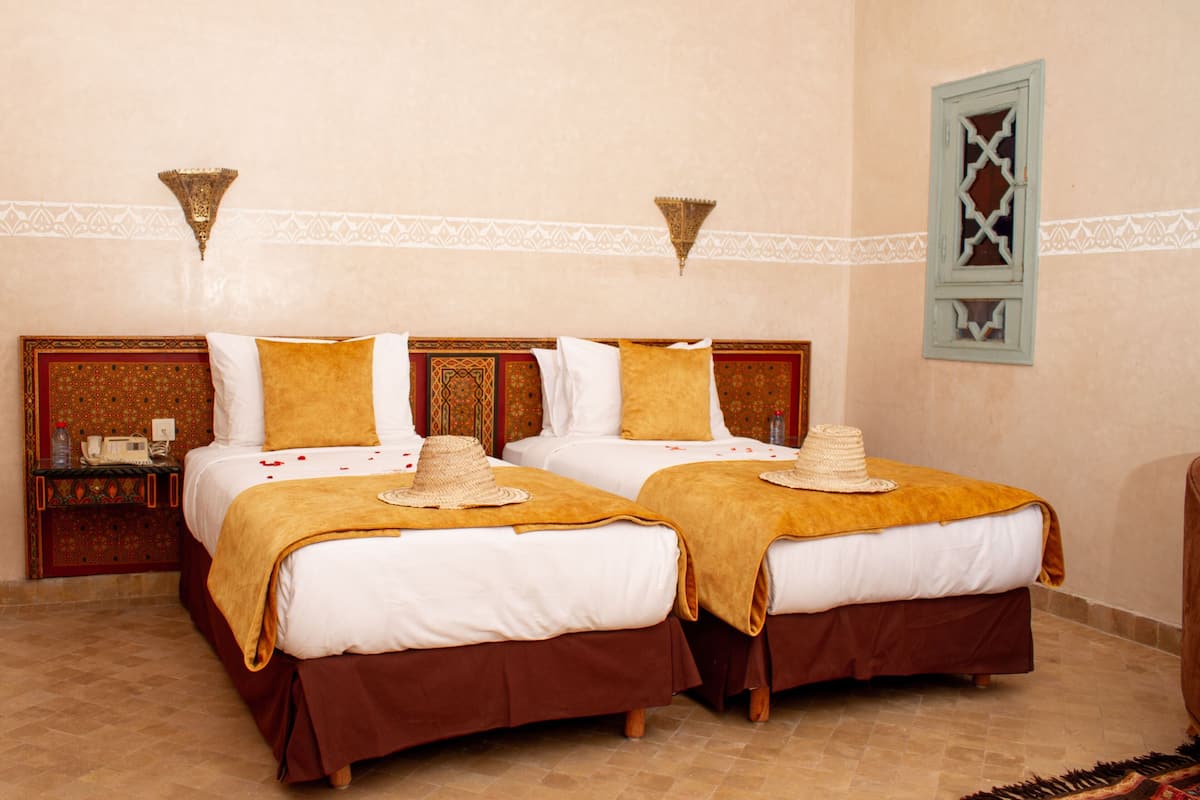 Una habitación de hotel con dos camas, cada una con una colcha marrón y ropa de cama blanca. Hay apliques decorativos y un tapiz con motivos en la pared, sobre las camas.