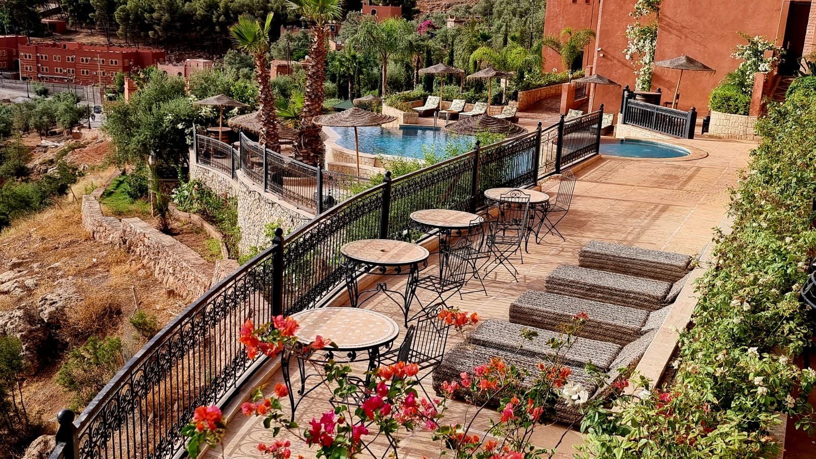 A rooftop pool area with lounge chairs and tables, surrounded by a metal railing and lush greenery.